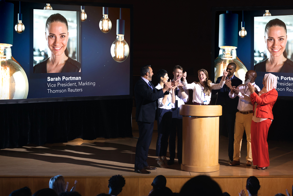 A group of models on stage pretending to be office workers accepting an award for excellence.