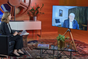 A webcasting live event with a woman seated in New York and a large flat screen showing a very old man talking